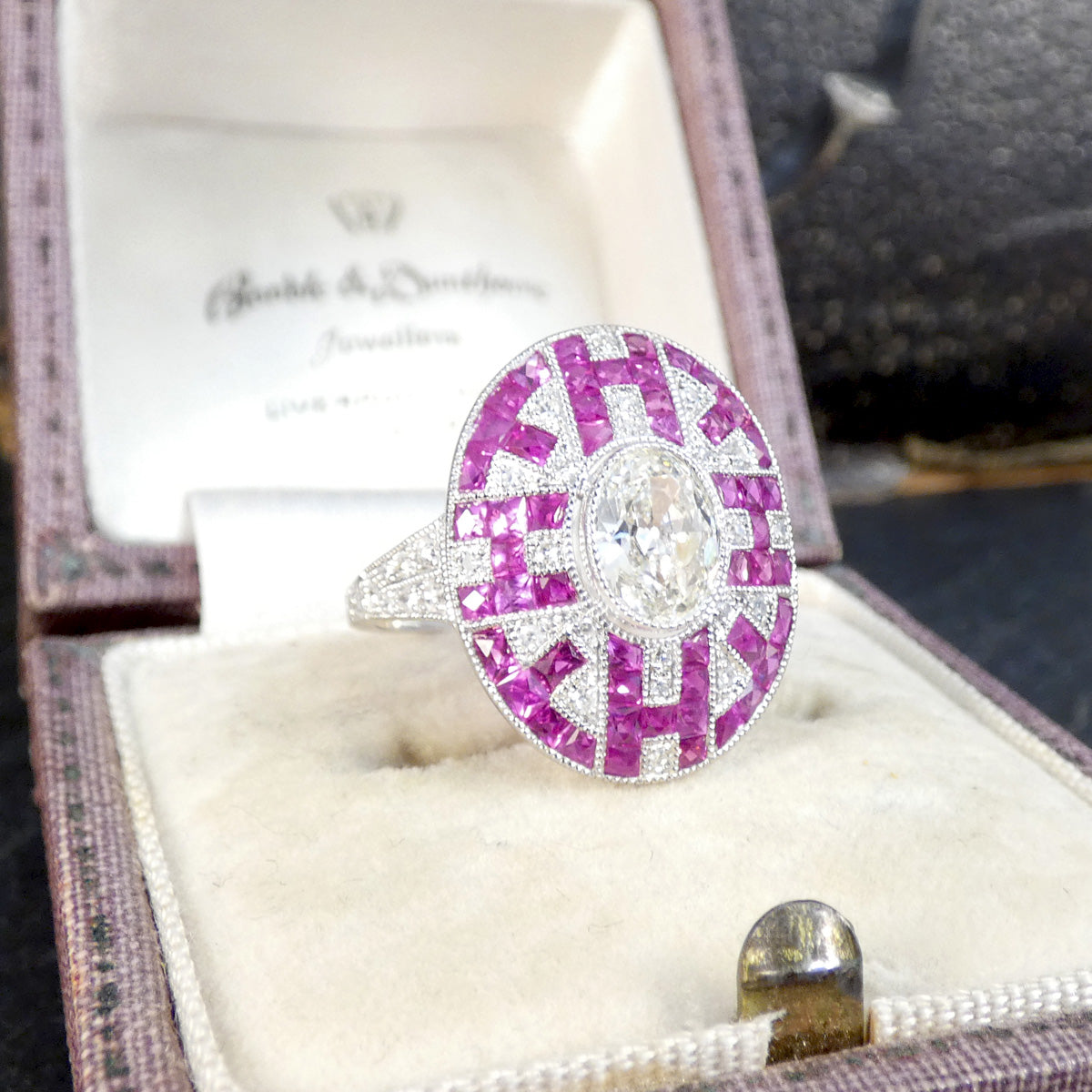 Art Deco Inspired Oval Cut Diamond Cluster Ring with Calibre Cut Rubies in Platinum displayed in a vintage box.