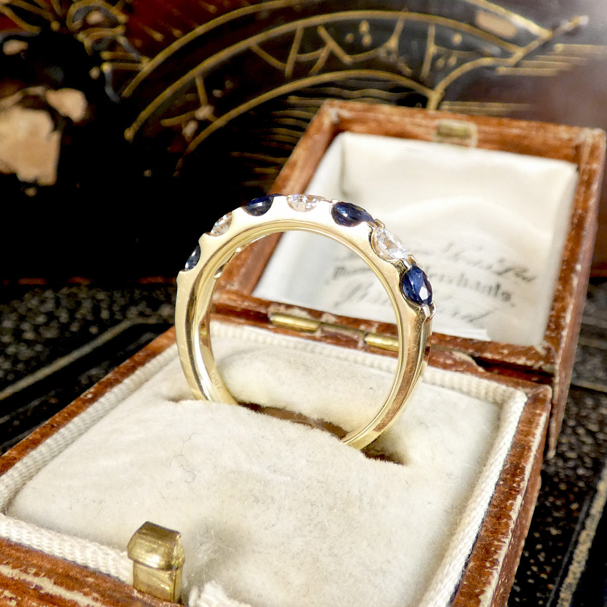 A yellow gold ring with dark blue sapphires and clear stones, displayed inside an open vintage wooden jewelry box with a cream velvet lining.