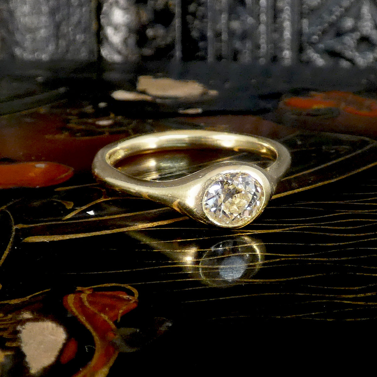 A gold ring with a round-cut clear gemstone in a bezel setting, resting on a glossy black decorative surface with a reflection below.