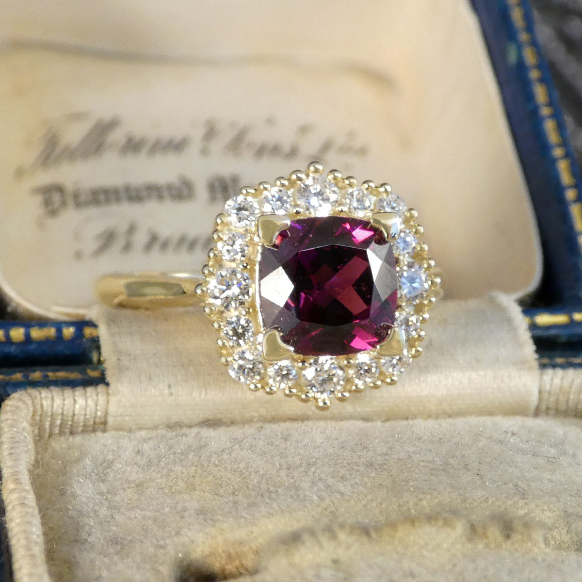 A vintage-style yellow gold ring featuring a cushion-cut deep magenta-purple center stone surrounded by a halo of small clear stones, displayed inside an open cream-colored jewelry box.