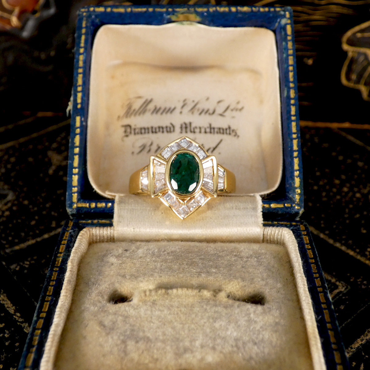 Elegant emerald and tapered diamond cluster ring set in yellow metal, testing as 18ct yellow gold, featuring a vivid oval emerald centre stone surrounded by shimmering tapered baguette diamonds, displayed in box.