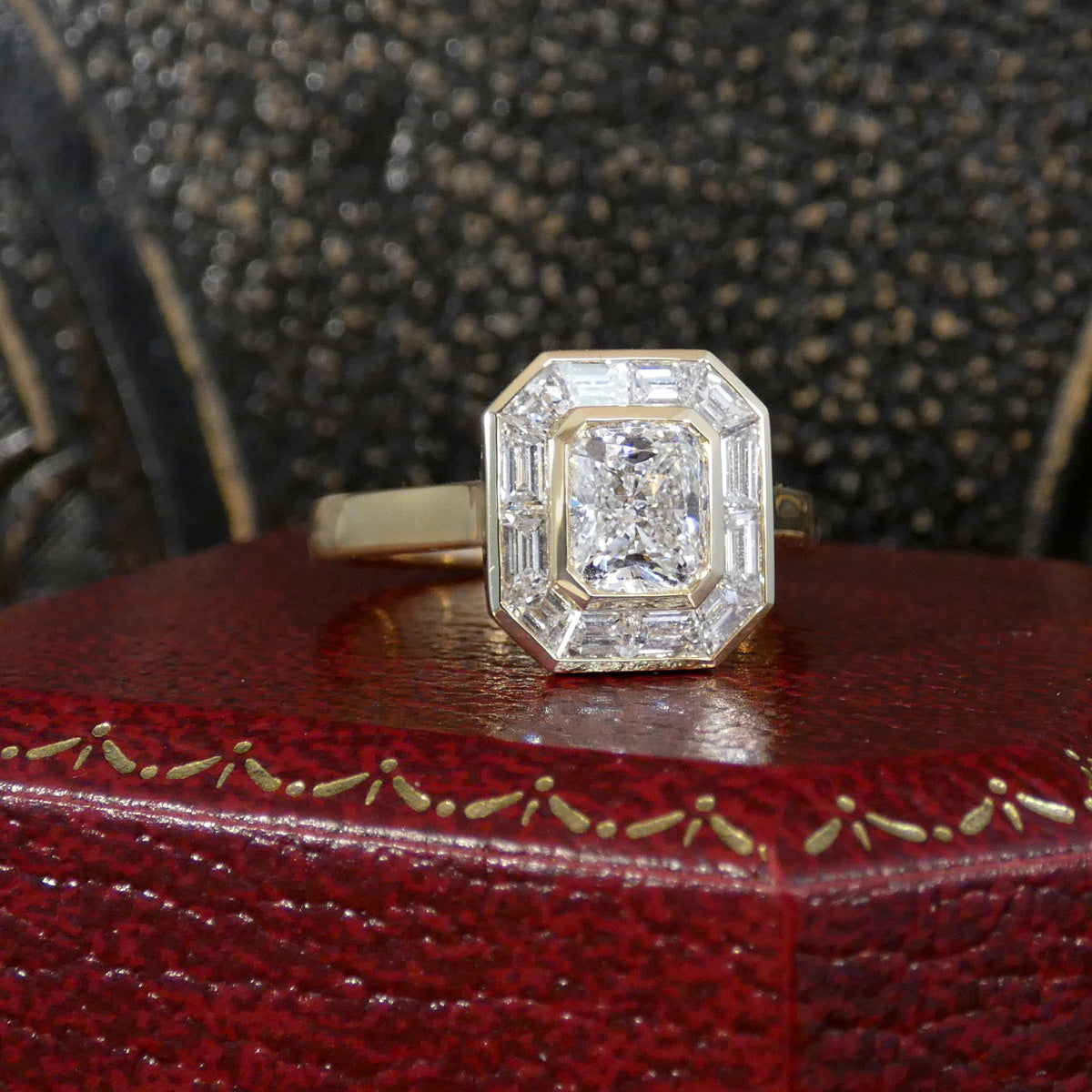 A gold ring with a large cushion-cut diamond surrounded by baguette-cut diamonds, displayed on a textured red box with a dark, sparkly background.
