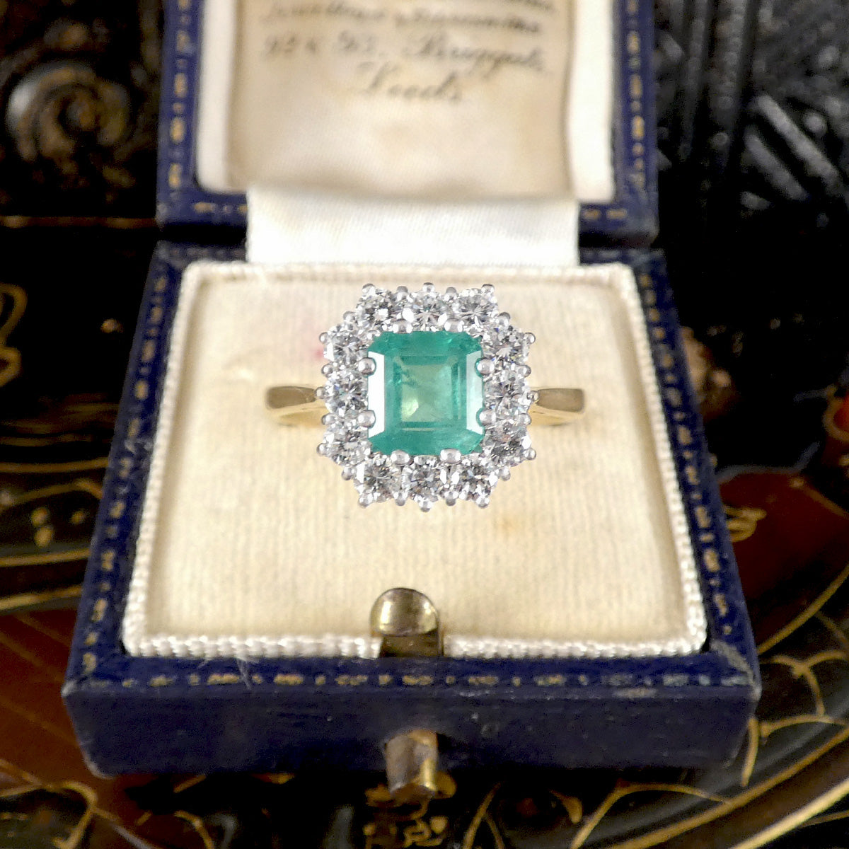 A vintage-style emerald-cut green gemstone ring with a halo of small clear stones, displayed inside an open dark blue velvet ring box.