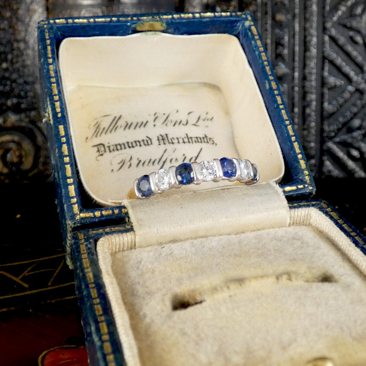 A vintage diamond and sapphire ring displayed inside an open blue velvet jewelry box with gold trim. The ring features alternating blue sapphires and clear diamonds set in silver-colored metal.