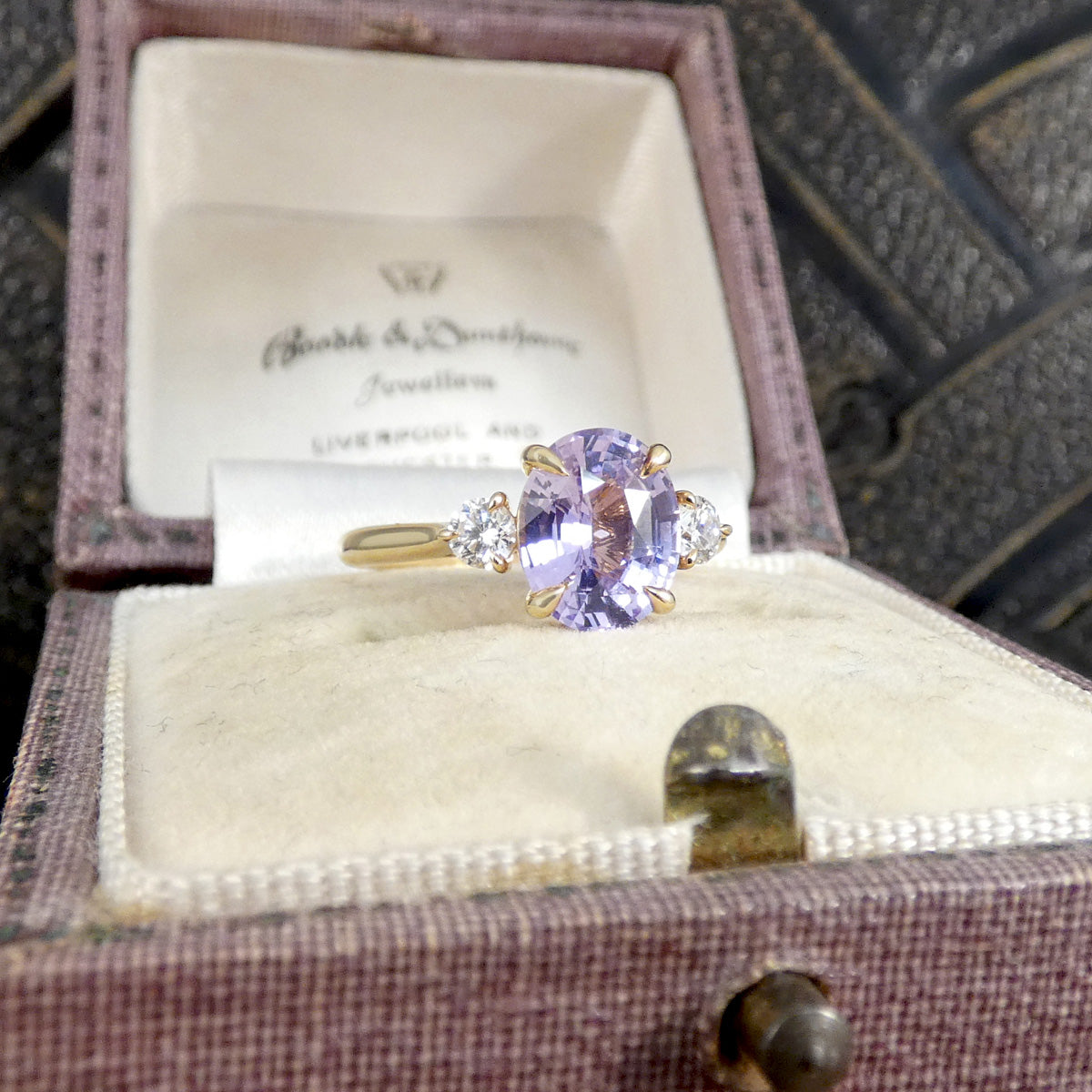 A yellow gold ring with a round-cut light purple center stone and small clear side stones, displayed inside an open vintage jewelry box. The inside lid shows the brand name 'Gable & Donaldson Jewellers'.