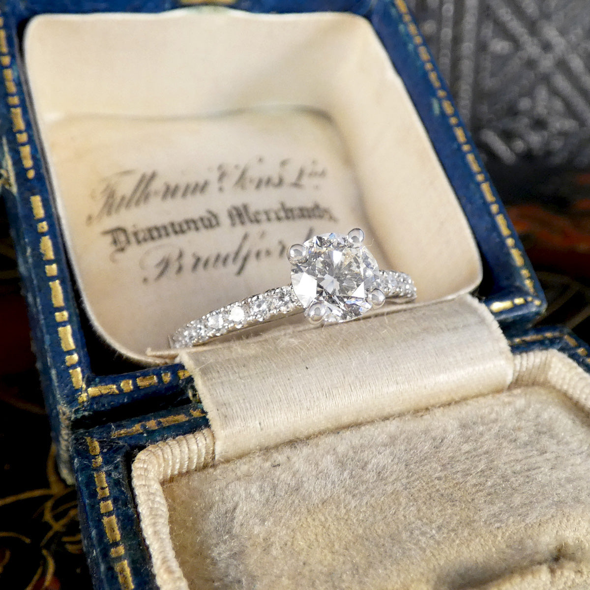 A diamond engagement ring with a round-cut center stone and a diamond-set band, displayed inside an open vintage blue velvet ring box with gold trim and cream lining.