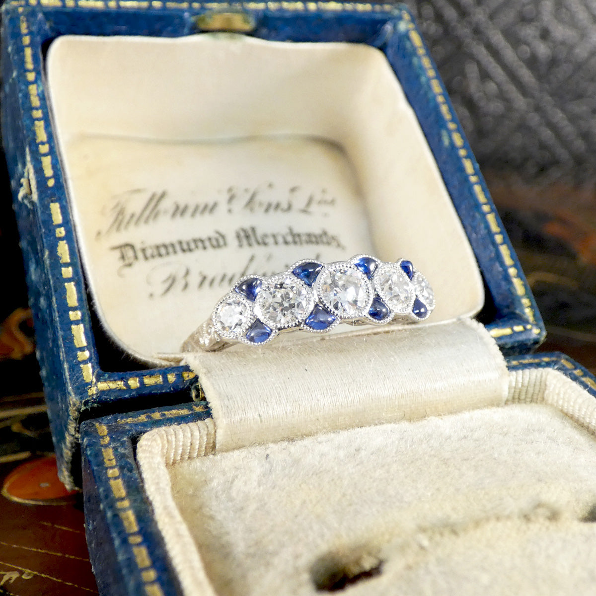 Angled view of Art Deco-style ring featuring old cut diamonds and calibre-cut sapphires set in platinum, presented in an antique blue velvet ring box with gold detailing.
