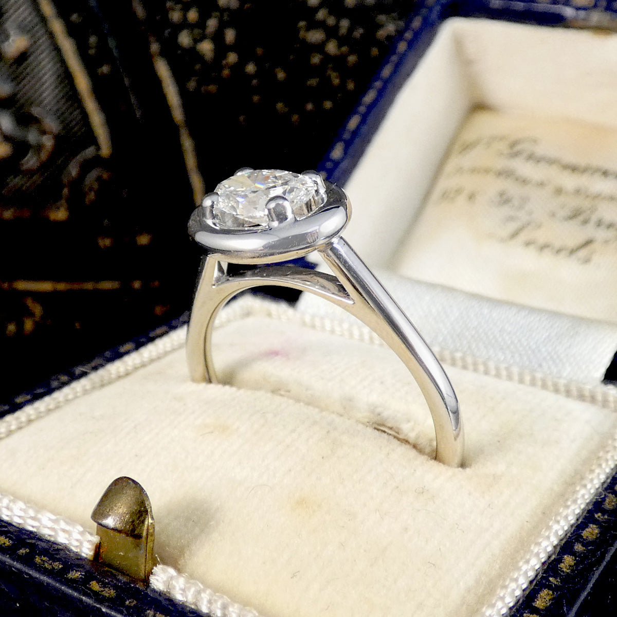 A silver solitaire ring with a round-cut diamond set in a vintage-style presentation box with a cream velvet cushion and a dark blue exterior.