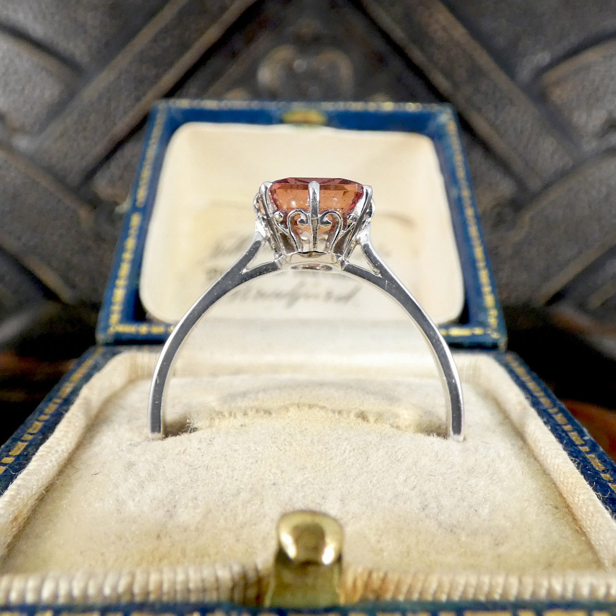 A silver ring with a round peach-colored gemstone in a crown-like setting, displayed inside an open vintage blue and cream jewelry box.