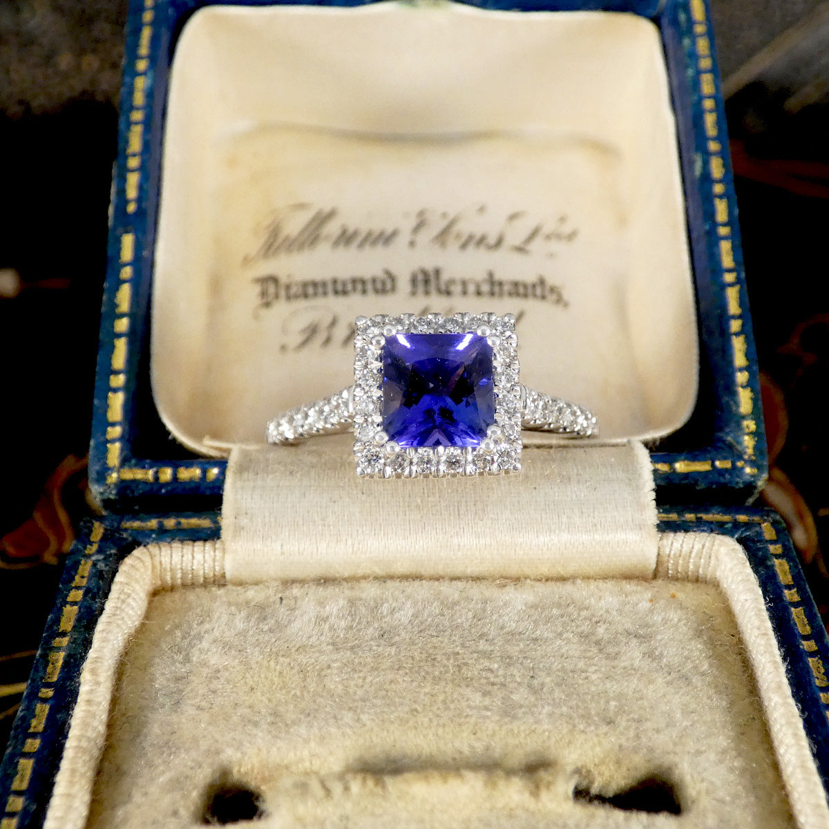 Diamond ring with a blue gemstone in an open jewelry box labeled 'Diamond Merchants'.
