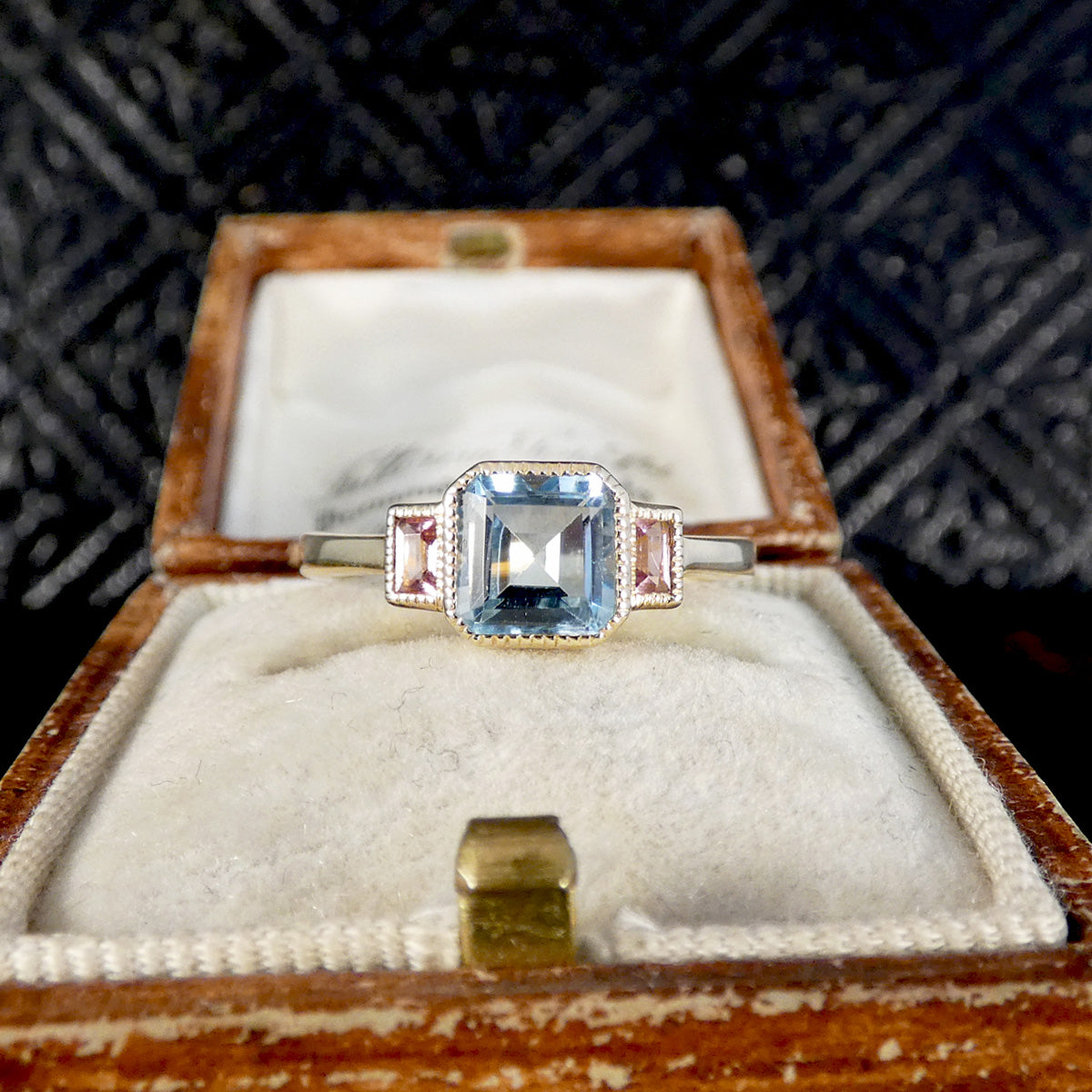 A vintage-style ring with a square-cut light blue center stone and small pink side stones, displayed in an open wooden jewelry box with a cream velvet interior.