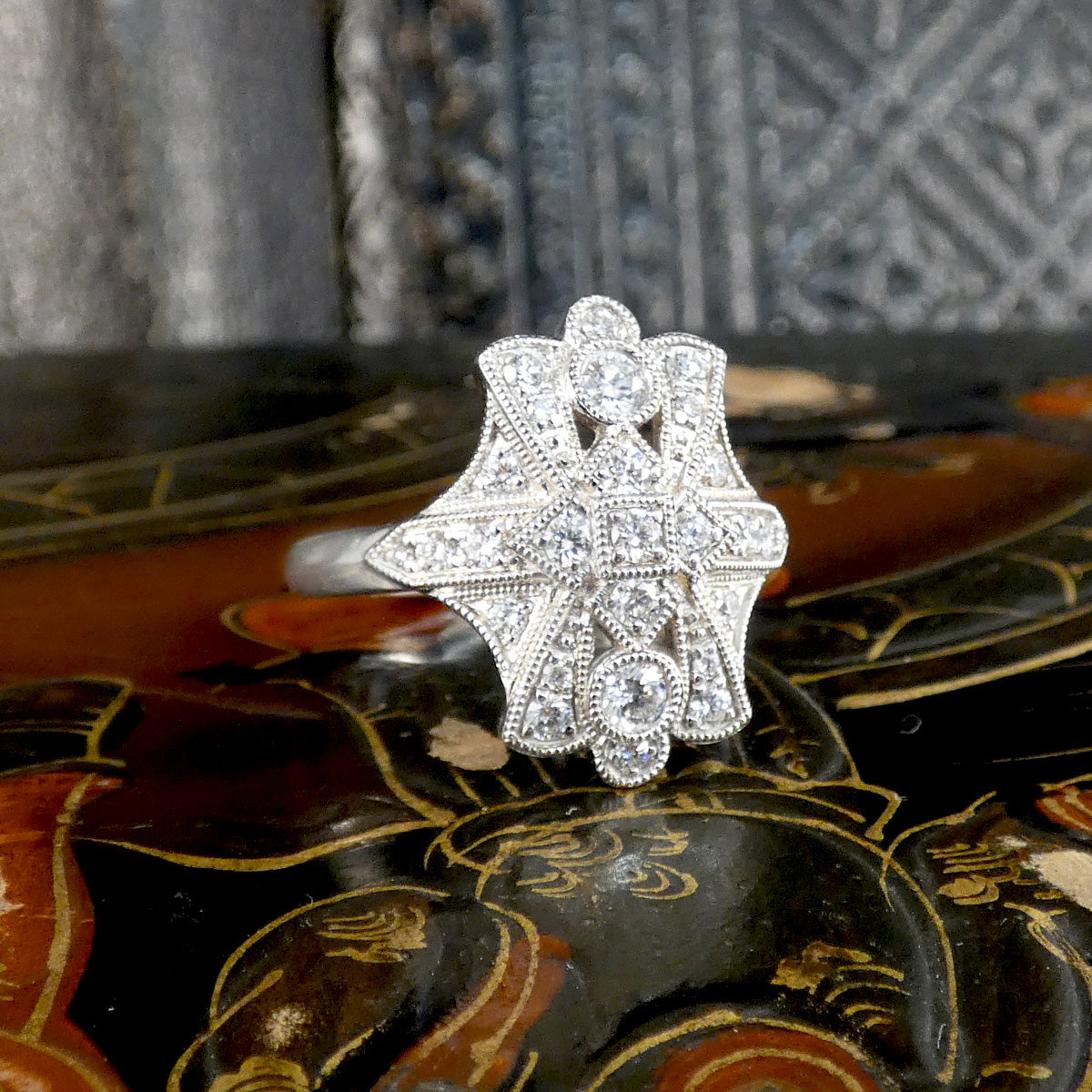 A silver-toned Art Deco style ring with milgrain detailing and clear gemstones, displayed on a dark patterned surface.