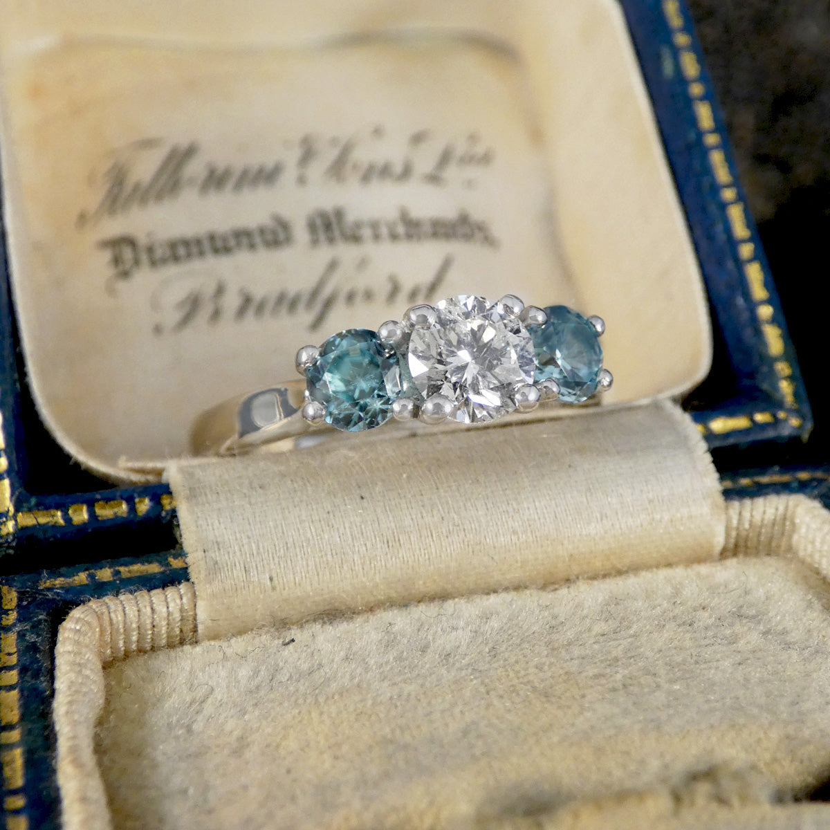 A vintage three-stone ring with round blue zircon side stones and a central round diamond, displayed inside an open antique ring box with cream velvet lining and a blue-gold exterior.