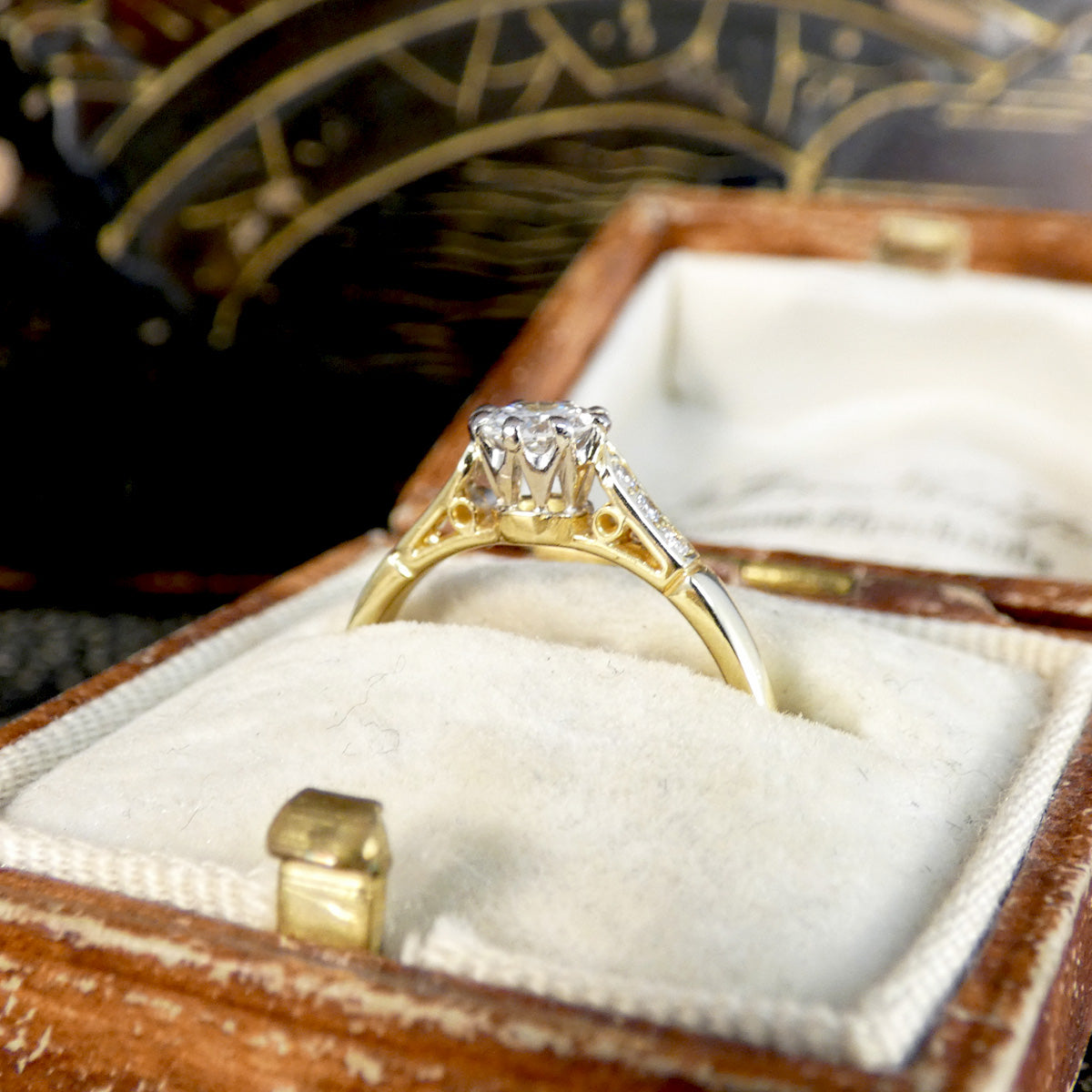 A yellow gold diamond ring with a round-cut center stone and diamond accents on the shoulders, displayed in an open vintage wooden ring box with a cream velvet insert.