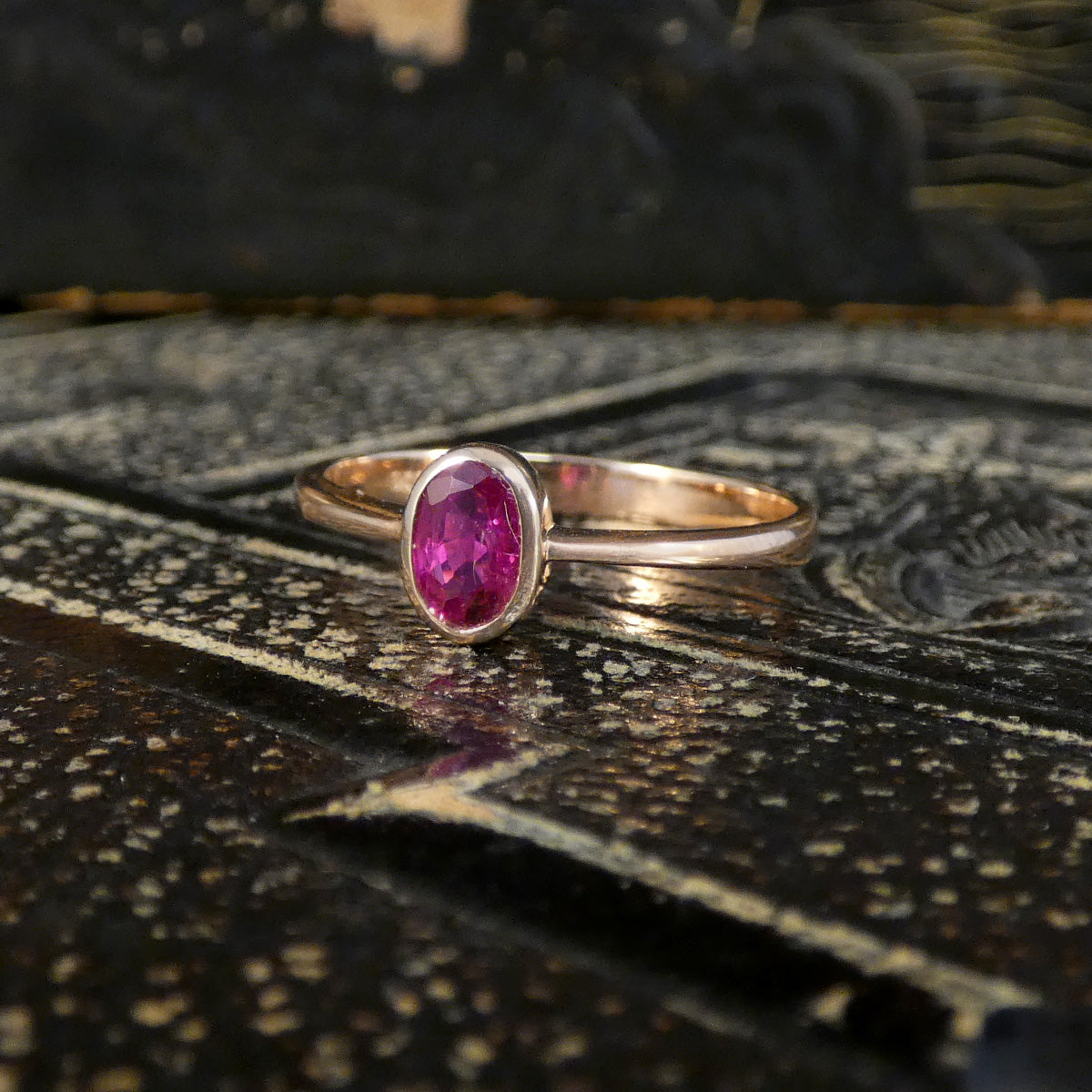 A rose gold ring with an oval-cut pink gemstone, resting on a dark, textured metallic surface.