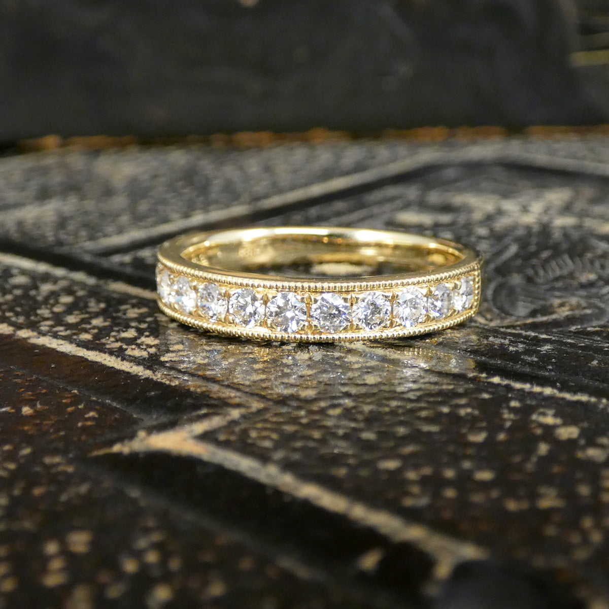 A yellow gold eternity ring with a row of round-cut clear gemstones, displayed on a dark, textured surface.