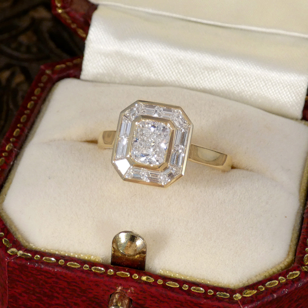A yellow gold ring with a large octagonal diamond and baguette-cut side stones, displayed in an open red and gold jewelry box with a cream satin lining.